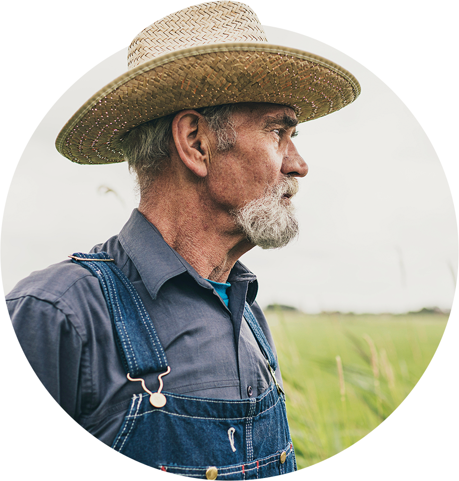 Side profile portrait of an older male farm worker wearing a straw hat, grey shirt, and blue overalls in front of a field looking into the distance to the right.